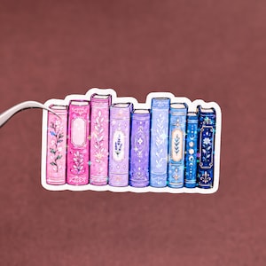 May include: A sticker featuring a row of nine illustrated books with spines in shades of pink, purple, and blue. The book spines are decorated with floral patterns, geometric shapes, and celestial motifs, accented with small, sparkling star details.