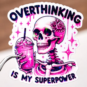 May include: Pink and black sticker with a glittery skeleton holding a pink drink. The sticker says "Overthinking is my superpower".