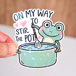 May include: A holographic sticker featuring a cartoon frog stirring a pot. The sticker has the text "ON MY WAY TO STIR THE POT" in white. The frog is light green with pink cheeks and is holding a wooden spoon.