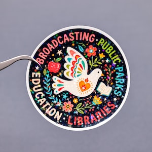 May include: Round sticker with a dove illustration, surrounded by text that reads "Broadcasting Public Parks Education Libraries." The dove has colorful wings and a heart-shaped design. The sticker has a dark background with stars and floral accents.