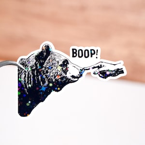 May include: A black and white sticker with a bear and a hand reaching out to touch its nose. The bear has a starry pattern and the hand is reaching out with the text "BOOP!" above it.