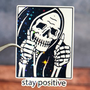 Puede incluir: Una pegatina en blanco y negro con un esqueleto que lleva una t&uacute;nica con capucha y da un pulgar hacia arriba con ambas manos. El fondo es un patr&oacute;n brillante en blanco y negro. El texto "stay positive" est&aacute; impreso debajo de la imagen.