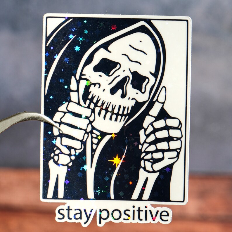 Puede incluir: Una pegatina en blanco y negro con un esqueleto que lleva una t&uacute;nica con capucha y da un pulgar hacia arriba con ambas manos. El fondo es un patr&oacute;n brillante en blanco y negro. El texto "stay positive" est&aacute; impreso debajo de la imagen.
