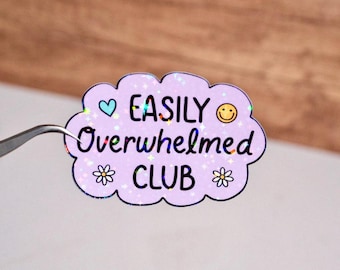 May include: A pink holographic sticker with the text "EASILY Overwhelmed CLUB" in black. The sticker has a heart, a smiley face, and two flowers.