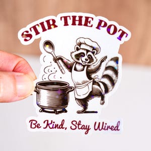 May include: A sticker featuring a raccoon chef holding a spoon over a pot. The text "STIR THE POT" arches above the raccoon, with "Be Kind, Stay Wired" below. The sticker has a holographic, glittery effect.