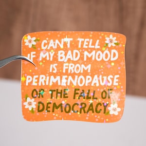 May include: Orange sticker with a glittery finish and white text that reads "Can't tell if my bad mood is from perimenopause or the fall of democracy".