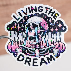 Puede incluir: Una pegatina holográfica con un esqueleto llorando bajo una nube oscura con estrellas. El texto "LIVING THE DREAM" está escrito alrededor del esqueleto.