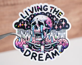 Puede incluir: Una pegatina holográfica con un esqueleto llorando bajo una nube oscura con estrellas. El texto "LIVING THE DREAM" está escrito alrededor del esqueleto.
