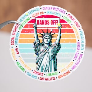 May include: A colorful circular sticker with a retro sunset design features the Statue of Liberty holding a sign that reads "HANDS OFF!"  The sticker is surrounded by a circle of text listing various social and political issues, including "Social Security", "Consumer", "Veteran Services", "Clean Air", "Immigrants", "Courts", "Public Lands", "Personal Data", "LGBTQ+ Rights", "Fair Elections", "Free Speech", "Schools", "Libraries", "Our Jobs", "Our Wallets", and "Our Books".