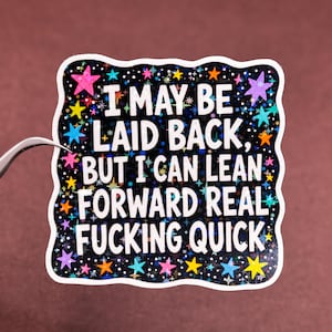 May include: A square sticker with a wavy white border featuring a black background filled with colorful, multi-sized stars. The white text reads: I MAY BE LAID BACK, BUT I CAN LEAN FORWARD REAL FUCKING QUICK. The design has a holographic finish.