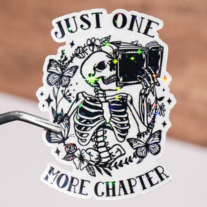 Puede incluir: Una pegatina holográfica con un esqueleto sosteniendo un libro y leyendo. La pegatina dice "Just One More Chapter".