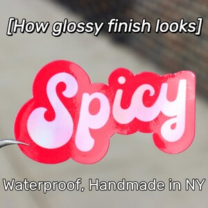 May include: A glossy red sticker with the word "Spicy" in white script. The sticker has a cloud-like shape and the text "Waterproof, Handmade in NY" is below the sticker.