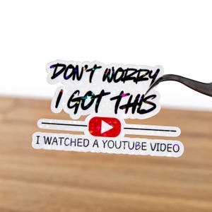 以下が含まれることがあります： 「DON'T WORRY I GOT THIS」と「I WATCHED A YOUTUBE VIDEO」の文字が入ったホログラフィックステッカー。ステッカーには赤いYouTube再生ボタンのアイコンが付いています。ピンセットでステッカーを持っています。