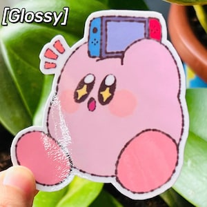 May include: A glossy sticker featuring a pink cartoon character holding a video game console. The character has large eyes with yellow star-shaped pupils, pink cheeks, and a small open mouth. The sticker has the word "Glossy" in the top left corner.