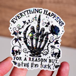May include: A holographic sticker with the text "EVERYTHING HAPPENS FOR A REASON BUT, what the fuck?". The sticker features a black skeleton hand giving the middle finger, a black rose, two skulls, and decorative vines.