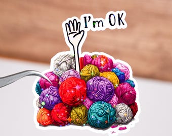 Op de afbeelding: Een sticker met een stapel kleurrijke bollen wol in tinten rood, paars, groen en geel. Een hand met de woorden "I'm OK" is boven de wol geheven. De sticker staat op een witte achtergrond.