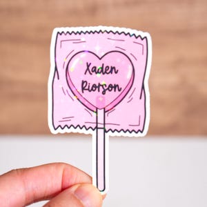 May include: A pink heart-shaped lollipop with the text "Xaden Riotson" inside. The lollipop is wrapped in a pink wrapper and is on a white stick.