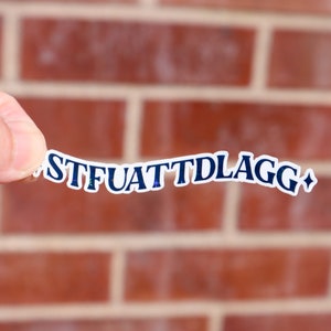 May include: A blue and white sticker with the text "STFUATT D LAGG" in a bold, stylized font. The sticker is curved and has a small diamond shape at the end.