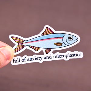 May include: A sticker featuring a cartoon fish with a light blue body, pink and yellow fins, and the text "full of anxiety and microplastics". The fish has a whimsical design with glitter accents.