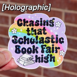 May include: Holographic sticker with the text "Chasing that Scholastic Book Fair high" in black, bubble letters. The sticker has a light purple cloud shape with a rainbow and flower accents. A stack of books is at the bottom.