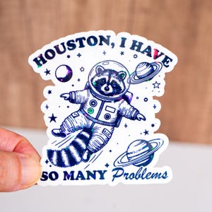 May include: A sticker featuring a raccoon in an astronaut suit, floating in space. The sticker has the text "Houston, I have so many problems" in a holographic font, along with planets and stars.