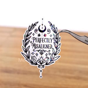 Op de afbeelding: Een witte sticker met de tekst "Perfectly Misaligned" in een decoratief lettertype, omgeven door een zwart bloemenontwerp met een halve maan en sterren. De sticker wordt vastgehouden door een pincet.