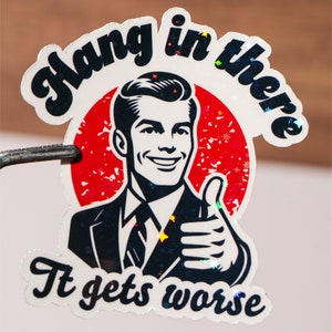 May include: A white sticker with a retro-style illustration of a man in a suit giving a thumbs up. The text on the sticker reads "Hang in there It gets worse".