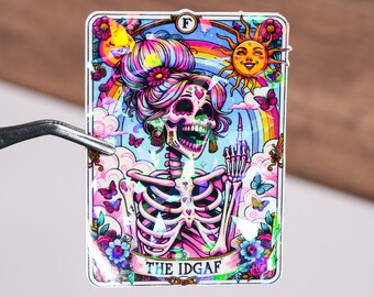 May include: A holographic sticker featuring a pink and white skeleton with a rainbow background and the text "THE IDGAF".