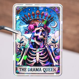 May include: A holographic sticker featuring a skeleton wearing a crown and floral wreath. The skeleton is laughing with its mouth open and its hands raised. The sticker has the text "THE DRAMA QUEEN" at the bottom.