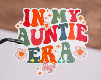 May include: A colorful sticker with the text "In My Auntie Era" in a retro font. The sticker features a butterfly, flowers, and glitter accents.