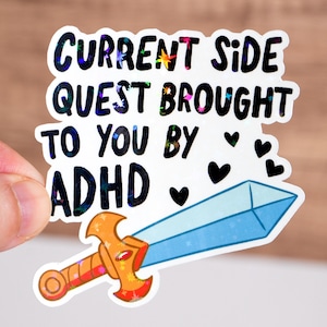 Op de afbeelding: Een witte sticker met een blauw en oranje zwaard en de tekst "CURRENT SIDE QUEST BROUGHT TO YOU BY ADHD" met hartjes rond de tekst.