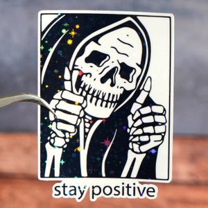 Puede incluir: Una pegatina en blanco y negro con un esqueleto que lleva una t&uacute;nica con capucha y levanta el pulgar con ambas manos. El fondo es un patr&oacute;n de purpurina negro e iridiscente. El texto "stay positive" est&aacute; impreso debajo de la imagen.