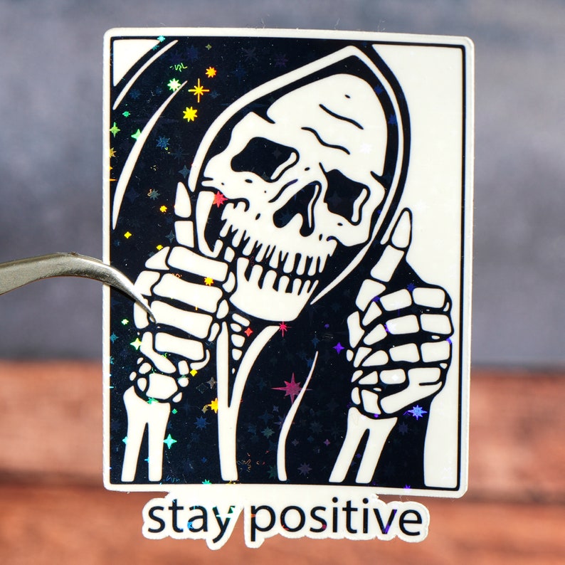 Puede incluir: Una pegatina en blanco y negro con un esqueleto que lleva una t&uacute;nica con capucha y levanta el pulgar con ambas manos. El fondo es un patr&oacute;n de purpurina negro e iridiscente. El texto "stay positive" est&aacute; impreso debajo de la imagen.