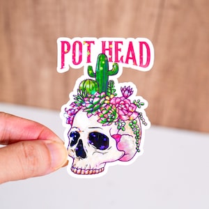 May include: A sticker depicting a skull adorned with a floral crown and a cactus. The pink text "POT HEAD" arches above the skull. The skull has purple and blue detailing. The sticker is held by a hand.