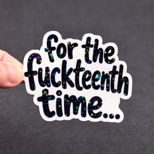 May include: A white sticker with the phrase "for the fuckteenth time..." in black, holographic lettering. The sticker has a glittery, star-like pattern within the letters and around the edges. The sticker is held against a dark gray background.