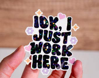 May include: A white sticker with a black and iridescent glitter design that says "IDK, I JUST WORK HERE". The sticker has pink and purple hearts and flowers around the text.