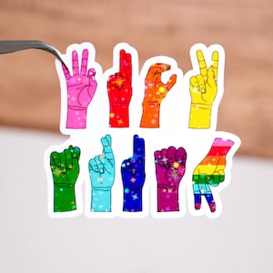 Puede incluir: Un conjunto de ocho pegatinas de mano de color arcoíris que representan el alfabeto del lenguaje de señas español. Las pegatinas están sobre un fondo blanco.