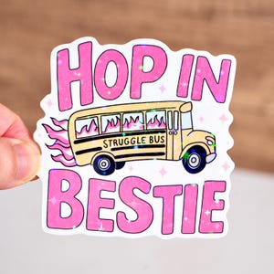 May include: A sticker with the text "HOP IN BESTIE" in pink, holographic lettering. A yellow school bus with flames and the words "STRUGGLE BUS" is in the center. The sticker has a white border and star accents.