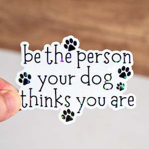 Może przedstawiać: Biała naklejka z czarnym tekstem, który brzmi "be the person your dog thinks you are". Naklejka jest ozdobiona kilkoma czarnymi śladami łap.