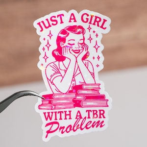 May include: A pink and white sticker with a cartoon illustration of a woman with her chin resting on her hands, smiling and looking down at a stack of books. The text "Just a girl with a TBR problem" is below the illustration.