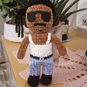 Könnte beinhalten: Eine handgefertigte Häkelpuppe mit braunem Garnkörper, schwarzem Haar mit seitlichen Dreadlocks und einer schwarzen Sonnenbrille aus Filz. Die Puppe trägt ein weißes Tanktop, eine hellblaue Hose mit schwarzem Gürtel und schwarze Schuhe.