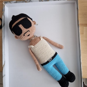 Könnte beinhalten: Eine handgefertigte Häkelpuppe mit gebräuntem Teint, schwarzem Haar und einer großen schwarzen Sonnenbrille. Die Puppe trägt ein weißes Unterhemd, eine silberfarbene Kettenhalskette, eine leuchtend blaue Hose mit schwarzem Gürtel und schwarze Schuhe.
