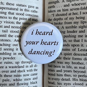 May include: A white button with black text that reads "i heard your hearts dancing!" The button is on top of a book with text.