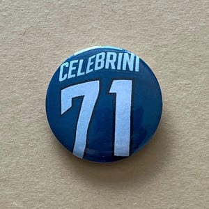 May include: A round, dark blue button with the name "CELEBRINI" arched across the top in white letters. Below, the number "71" is displayed in a light blue, bold font. The button has a glossy finish.