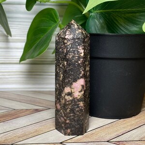 May include: A black and pink polished crystal point with a natural, rough texture. The crystal is shaped like a pyramid with a pointed top and a flat base.