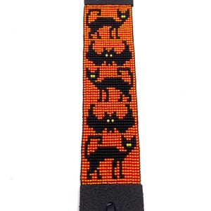 May include: A rectangular beaded bracelet featuring a vibrant orange background with a repeating pattern of black cats and bats. The black figures have small, bright yellow eyes. The beaded section is attached to black leather ends with black cords.