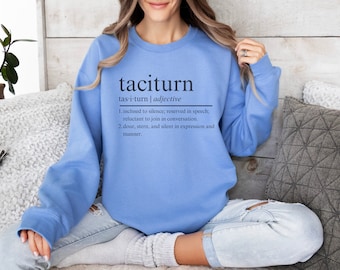 May include: A light blue sweatshirt with the word "taciturn" and its definition printed on the front. The definition is: "tas-i-turn | adjective 1. inclined to silence; reserved in speech; reluctant to join in conversation. 2. dour, stern, and silent in expression and manner."