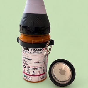 May include: An orange cylindrical container labelled PUFFTRACKR with a black cap and a light purple mouthpiece. A black ring with a metal key ring holds a circular silver Apple AirTag. The label includes text like PROXY CORE and 710 LIFE.