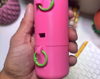 以下が含まれることがあります： マットな質感の鮮やかなピンク色の円筒形容器で、外側に2つのライムグリーンの円形リングが取り付けられています。上部と下部のセクションを分ける水平な継ぎ目があり、側面に小さな長方形の開口部があります。