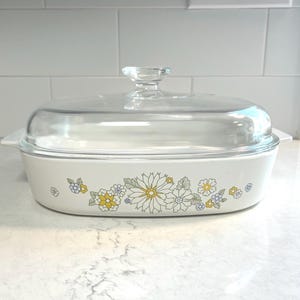 May include: A white casserole dish with a clear glass lid. The dish is decorated with a floral pattern in yellow, blue, and white. The dish is rectangular with rounded edges and has two small handles.